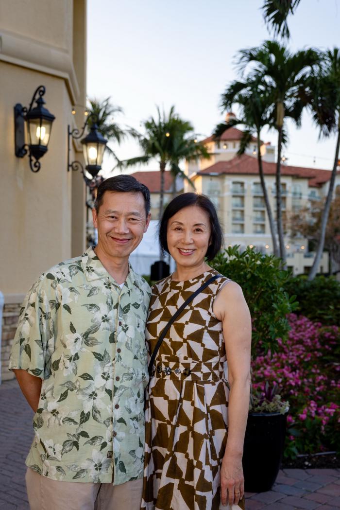Peter Li, left, and Jingli Yang, right, are standing side-by-side outdoors, smiling at the camera. The man is wearing a short-sleeved, light green Hawaiian shirt, and the woman is wearing a sleeveless dress with a brown and white geometric pattern. In the background are palm trees, tropical foliage, and a large resort-style building with a terracotta roof.