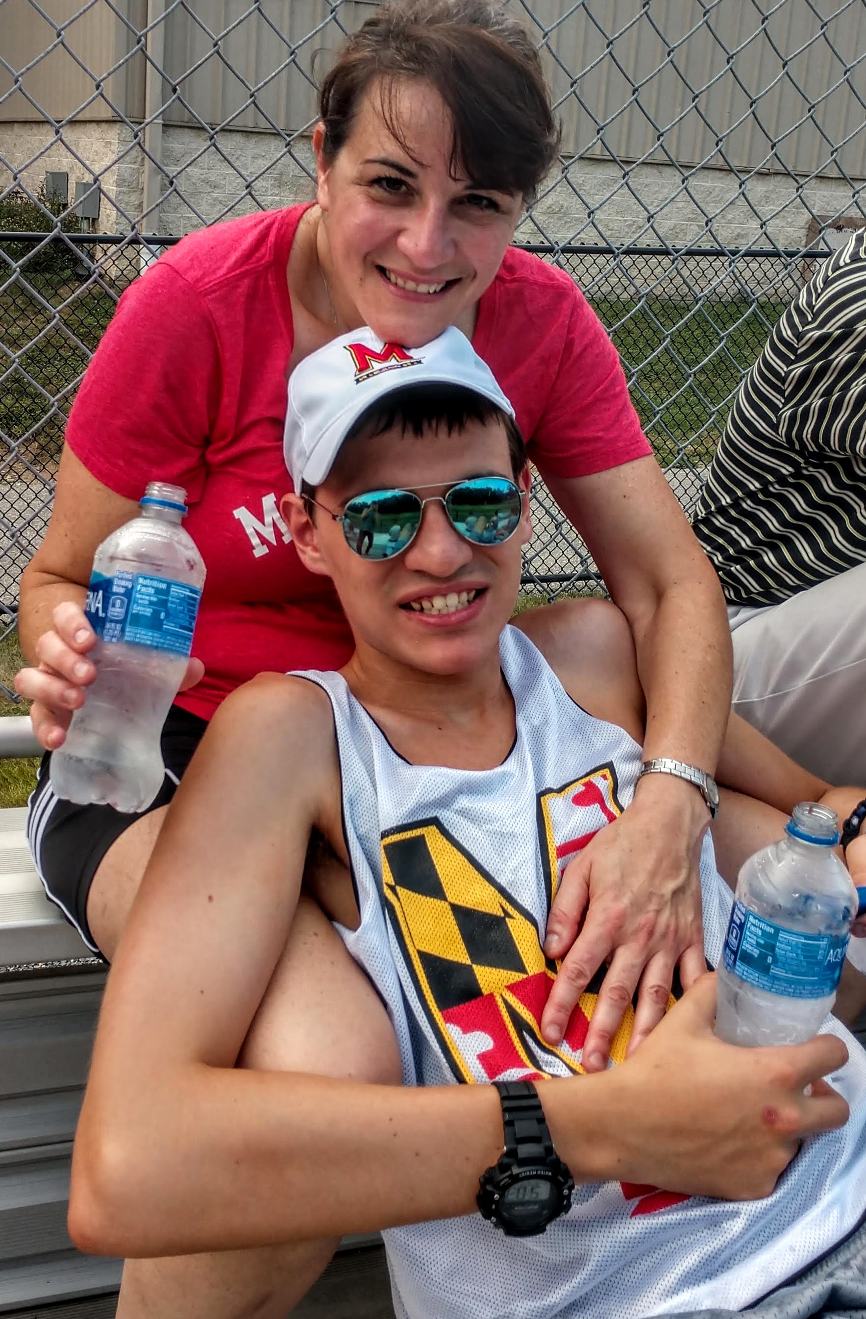 Lisa Wiederlight, in a red shirt, and her son Joshua, in a white University of Maryland jersey, sit together on bleachers, both smiling. Lisa leans her chin on Joshua's white baseball cap, and both hold plastic water bottles. They are in the bleachers watching a UMD field hockey game.