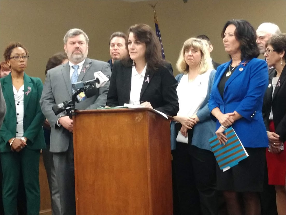 Lisa Wiederlight, with long dark hair in a black blazer, stands behind a wooden podium advocating for the passage of “Kevin and Avonte’s Law Act.” She is surrounded by a group of men and women in professional attire, several of whom are wearing small purple ribbons on their lapels.