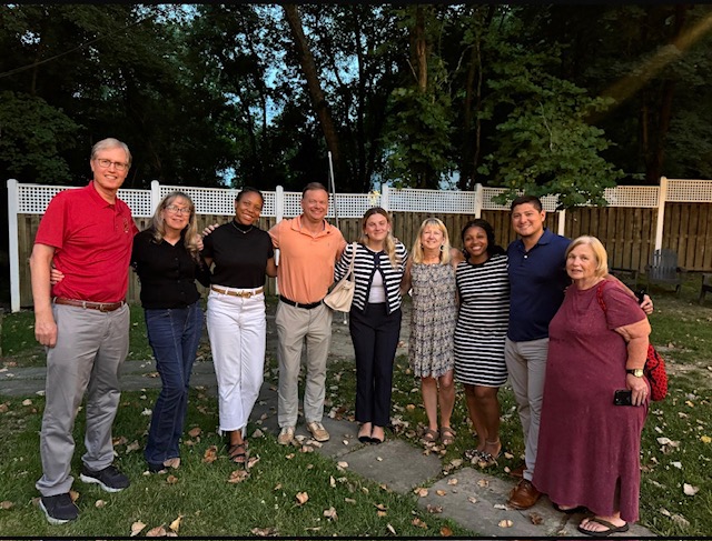 Photo of the Baltimore Terps Alumni Network leadership team, April Smith in a white and black striped dress the third from the right, standing in a backyard with green grass, leaves on the ground, and a wooden fence