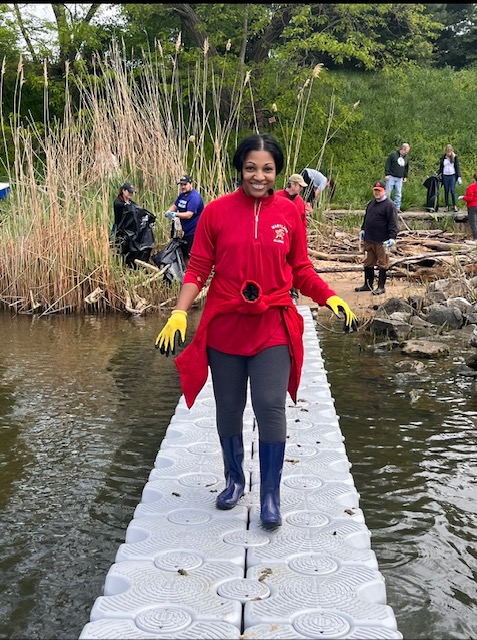 photo of April Smith standing on a bridge at a park clean up event wearing yellow gloves, and a red UMD quarter zip.