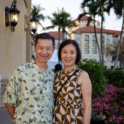 Peter Li, left, and Jingli Yang, right, are standing side-by-side outdoors, smiling at the camera. The man is wearing a short-sleeved, light green Hawaiian shirt, and the woman is wearing a sleeveless dress with a brown and white geometric pattern. In the background are palm trees, tropical foliage, and a large resort-style building with a terracotta roof.