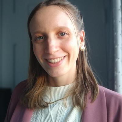 Headshot photo of Ren Salig smiling at the camera and wearing a white crewneck sweater, gold necklace and earrings, and a mauve blazer in front of a light grey background.