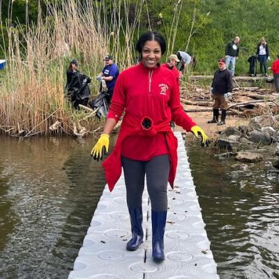 photo of April Smith standing on a bridge at a park clean up event wearing yellow gloves, and a red UMD quarter zip.
